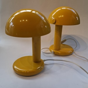 May include: Two matching table lamps with a glossy yellow finish. Each lamp features a rounded, dome-shaped shade supported by a cylindrical stem and a circular base. A white power cord extends from the base of each lamp, and a small knob is visible.