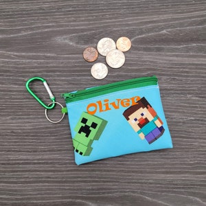 May include: A light blue coin purse with a green zip and a green carabiner clip. The purse features a green Creeper and a pixelated Steve character, with the name "Oliver" in orange. Several coins are scattered nearby.