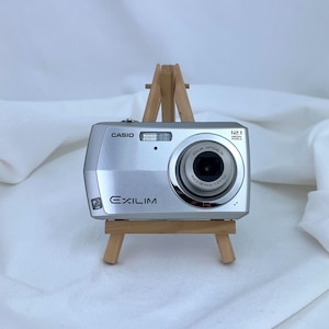 May include: A silver Casio Exilim digital camera with a 12.1 megapixel lens and built-in flash. The compact camera features a metallic finish, a circular lens barrel with technical specifications printed around it, and is displayed on a wooden easel.