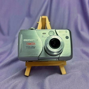 May include: A silver Olympus Camedia C-50 Zoom digital camera displayed on a miniature wooden easel. The camera features a black lens and the text "OLYMPUS CAMEDIA DIGITAL CAMERA C-50 ZOOM". The background is a purple fabric.