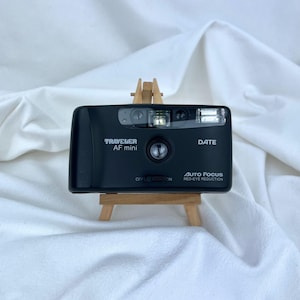 May include: A black Traveler AF mini 35mm film camera with a central lens, built-in flash, and red-eye reduction feature. The front body displays the text Traveler AF mini, DATE, and Auto Focus Red-Eye Reduction with an off and on power switch.