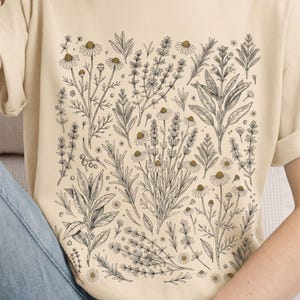 Soft Ivory cotton tee with a Victorian botanical scatter of kitchen herbs in dark charcoal ink, worn against pale denim — Scattered Kitchen Herbs Comfort Colors shirt for cottagecore witches and herb gardeners. By MythicThreadMagic.