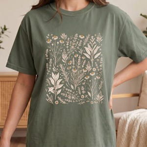 Sage Moss Comfort Colors tee photographed from a side angle in a botanical studio setting with dried flowers and natural wood, showing the relaxed drape of the Scattered Kitchen Herbs shirt for forest witches. By MythicThreadMagic.