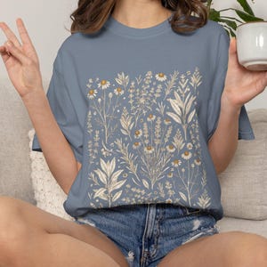 Faded Blue Jean Comfort Colors tee with cream botanical herb print, model holding a matching Kitchen Witch Herbs ceramic mug in a sunlit corner — relaxed cottagecore styling for tea-loving home apothecaries. By MythicThreadMagic.