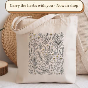 A cream-colored canvas tote bag featuring a detailed black and white botanical illustration of various herbs, including chamomile, lavender, rosemary, and sage, with subtle hints of yellow. The tote is displayed in a cozy, neutral-toned setting with a potted aloe vera plant, woven mats, and a textured pillow. A banner at the top reads, "Carry the herbs with you - Now in shop.