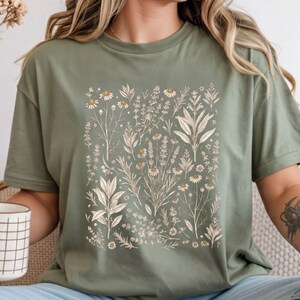 Sage Moss green cotton tee featuring a scattered botanical specimen print of kitchen herbs in cream ink, styled with dried grasses and a terracotta vessel in a warm cottagecore study. By MythicThreadMagic.