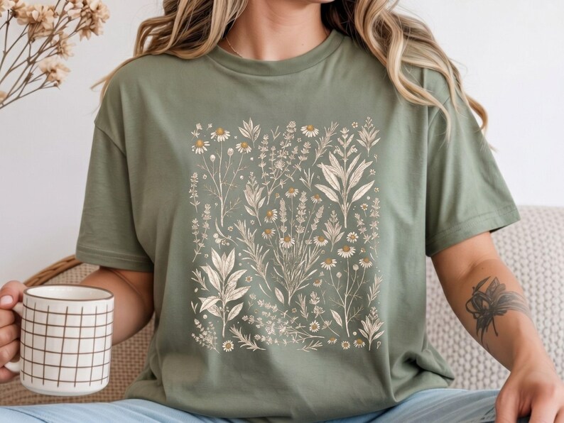 Sage Moss green cotton tee featuring a scattered botanical specimen print of kitchen herbs in cream ink, styled with dried grasses and a terracotta vessel in a warm cottagecore study. By MythicThreadMagic.
