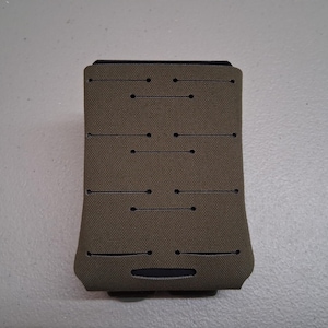 May include: A tan-colored, rectangular pouch with multiple horizontal slits and circular perforations. The pouch has a black base and a black top. The surface has a textured appearance, and the design suggests a tactical or organizational purpose.