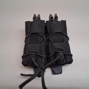 May include: Black tactical magazine pouch. The pouch is made of durable nylon fabric and features two compartments. Each compartment has a retention system and bungee cords for secure storage. The pouch is designed to hold magazines and other tactical gear.