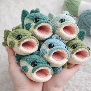 May include: A collection of handmade crochet fish keychains in various shades of green and blue. Each fish has a wide-open mouth with a pink interior, black eyes, and a keychain attachment. The fish are held in a hand.