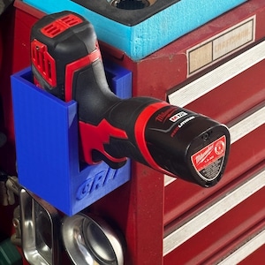 May include: A blue 3D-printed holder with the word "GRIP" on the side, designed to hold a black and red Milwaukee M12 cordless drill. The drill is mounted on a red toolbox with a Craftsman label.