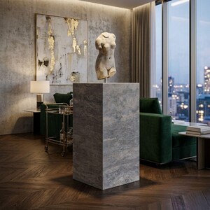 May include: A classical torso sculpture is displayed on a grey stone plinth. The plinth is rectangular and stands on a dark wood floor. The background includes a large abstract painting with gold accents and a green sofa.
