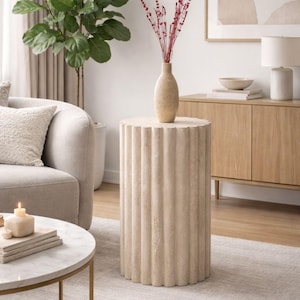 May include: A beige, cylindrical side table with vertical fluted detailing. A beige vase with red stems sits atop the table. The table is in a living room setting with a sofa, rug, and other furniture.