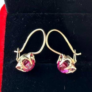 May include: A pair of gold-toned earrings featuring round, faceted, vibrant pink stones set in a four-prong metal basket setting. The earrings are designed with curved wire hooks that extend upward from the base of the stone setting.
