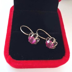 Könnte beinhalten: Ein Paar silberfarbene Ohrringe mit runden, facettierten, leuchtend pinken Steinen in einer gewellten Metallfassung. Die Ohrringe haben gebogene Haken und liegen in einer kleinen, rechteckigen Schmuckschatulle mit rotem Rand und schwarzem Inlay.