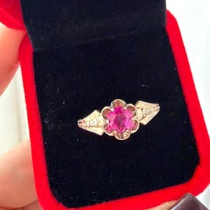 May include: A silver-toned ring featuring a central, vibrant pink, six-pointed star-shaped gemstone set within a flower-shaped metal frame. The band displays a series of small, circular indentations along the sides of the setting.