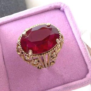 May include: A gold ring featuring a large, oval-cut, deep red gemstone held by four prongs. The band and setting are crafted with an intricate, openwork filigree design, showcasing a textured, rope-like metal pattern around the central stone.