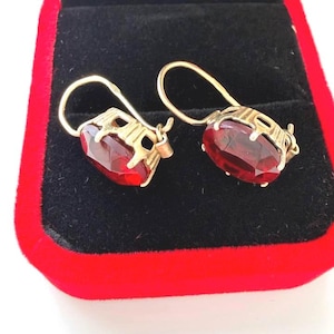 May include: A pair of gold earrings featuring oval-cut deep red gemstones. Each earring has a secure hook-style closure and a decorative metal setting that holds the faceted stone in place, showcasing a classic design with a polished finish.