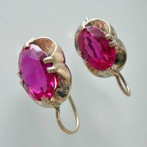 May include: A pair of vintage silver earrings featuring large, oval-cut, magenta-colored gemstones. The earrings have a simple, elegant design with a hook closure. The gemstones are set in a silver metal setting with a decorative edge.