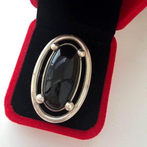 May include: A silver brooch featuring a large, smooth, oval-shaped black stone set within a polished silver frame. The frame is composed of two concentric oval bands connected by four small silver spheres positioned at the top and bottom.