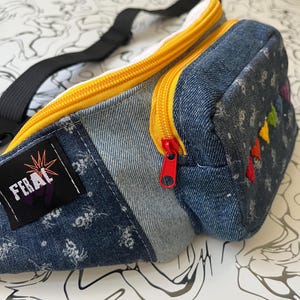 Puede incluir: Una riñonera de mezclilla con una etiqueta negra que muestra el texto FERAL y un gráfico de estrella. El bolso tiene una cremallera amarilla brillante con un tirador rojo, una correa negra ajustable y un pequeño patrón de triángulos arcoíris bordado.