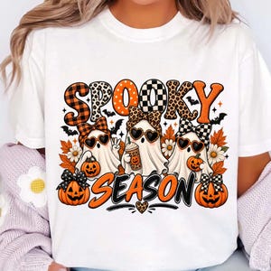 Puede incluir: Una camiseta blanca con un gráfico que incluye el texto SPOOKY SEASON en patrones naranjas y negros. El diseño muestra tres fantasmas con gafas de sol en forma de corazón y lazos estampados, rodeados de calabazas, hojas de otoño y murciélagos.