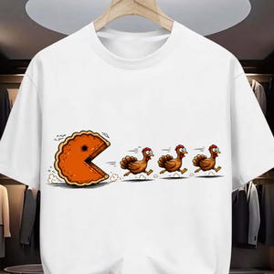 May include: A white short-sleeve t-shirt featuring a graphic print of a large orange pie slice shaped like a video game character chasing three brown turkeys across the front of the garment. The design includes motion lines and dust clouds.