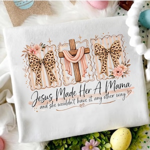 May include: White t-shirt featuring the text "Jesus Made Her A Mama" and decorative elements. The design includes a wooden cross with pink fabric, leopard print bows, and floral accents. The t-shirt has a crew neck.
