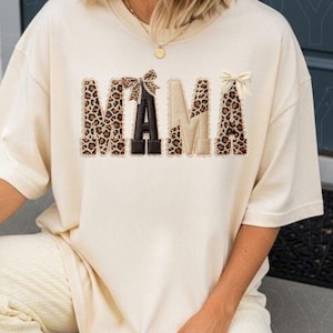 May include: Cream-colored sweatshirt with the word "MAMA" in a decorative design. The letters are a mix of leopard print, black, and tan, with small bows on top. The sweatshirt has a relaxed fit.