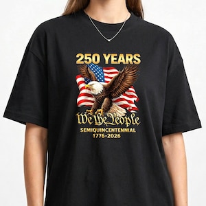 Könnte beinhalten: Schwarzes T-Shirt mit einer Grafik der amerikanischen Flagge und einem Weißkopfseeadler. Der Text auf dem Shirt lautet "250 YEARS" über der Flagge und "We the People" darunter, mit "SEMIQUINCENTENNIAL 1776-2026" am unteren Rand.