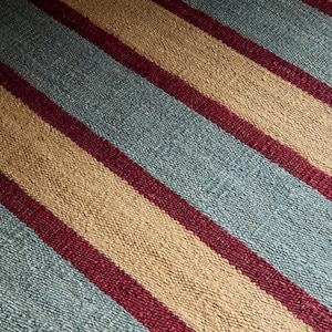 May include: Close-up of a woven textile with a striped pattern. The design features alternating bands of light blue, tan, and burgundy. The texture appears coarse and the stripes run diagonally across the frame.