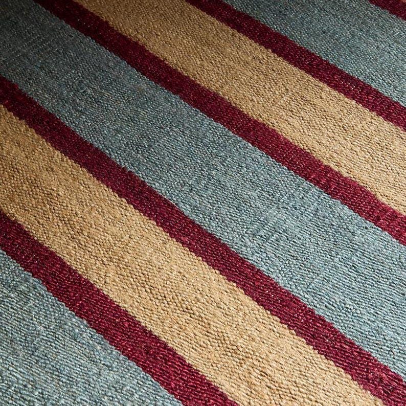 May include: Close-up of a woven textile with a striped pattern. The design features alternating bands of light blue, tan, and burgundy. The texture appears coarse and the stripes run diagonally across the frame.