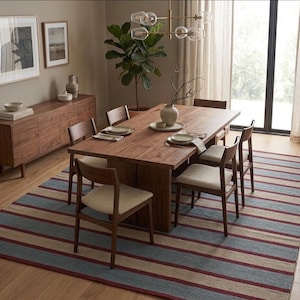 May include: A dining room scene featuring a large wooden table with six chairs, set on a striped rug. The rug has blue, beige, and red stripes. A wooden sideboard and framed artwork complete the room's decor.
