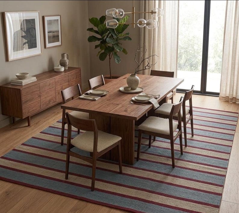 May include: A dining room scene featuring a large wooden table with six chairs, set on a striped rug. The rug has blue, beige, and red stripes. A wooden sideboard and framed artwork complete the room's decor.