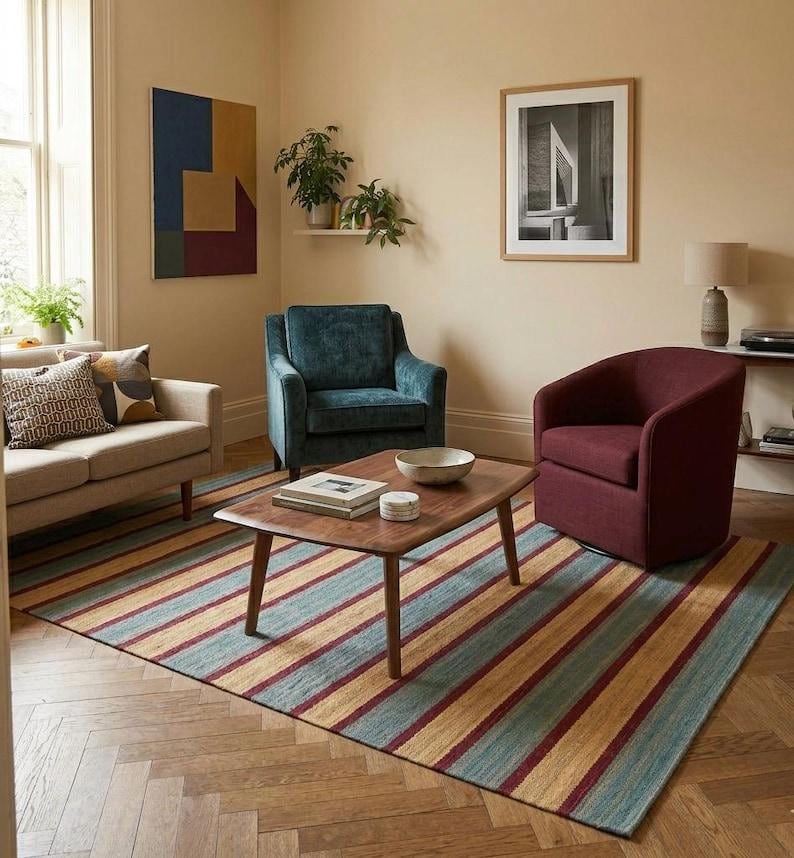 May include: A living room scene featuring a beige sofa, teal armchair, and burgundy swivel chair. A striped rug with teal, gold, and burgundy stripes sits beneath a wooden coffee table. Artwork and plants add decorative elements to the room.