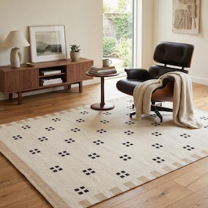 Op de afbeelding: Een rechthoekig vloerkleed met een crèmekleurige basis, een herhalend patroon van kleine zwarte ruitvormen en een brede, getextureerde beige rand. Het kleed ligt op een houten vloer en is gedeeltelijk bedekt door een bruine leren fauteuil.