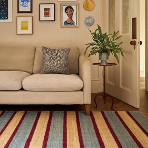 May include: A beige sofa with a leopard print pillow sits in front of a wall with framed artwork. A red, blue, and tan striped rug covers the floor. A potted plant on a small red stand is next to the sofa. The room has a neutral color palette.