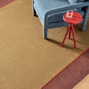 Könnte beinhalten: Ein blauer Polstersessel steht auf einem großen Teppich mit einem breiten senfgelben Mittelteil, der von dunkelroten Abschnitten begrenzt wird. Ein kleiner, runder, leuchtend roter Dreibein-Beistelltisch trägt ein klares Wasserglas.