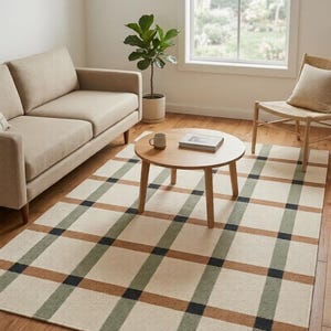 May include: A rectangular area rug featuring a large-scale grid pattern in shades of cream, sage green, tan, and black. The rug has a flat-woven texture and covers a portion of the wooden floor, extending underneath a round wooden coffee table.