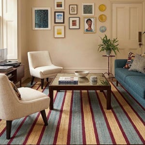 May include: A living room scene featuring a blue sofa, two cream-colored armchairs, and a striped rug with red, blue, and tan stripes. Artwork hangs on the wall above a dark wood coffee table. A plant sits on a small table.