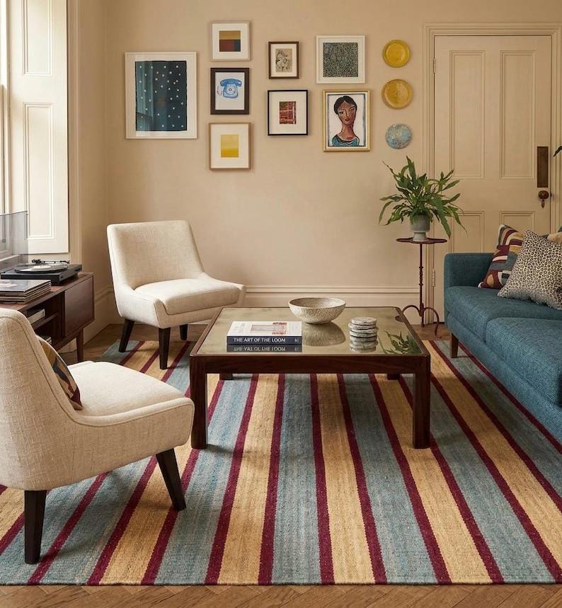 May include: A living room scene featuring a blue sofa, two cream-colored armchairs, and a striped rug with red, blue, and tan stripes. Artwork hangs on the wall above a dark wood coffee table. A plant sits on a small table.