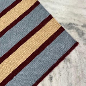 May include: A close-up of a striped rug with alternating bands of light blue, beige, and burgundy. The rug's texture is visible, and the color scheme creates a visually appealing pattern. The rug is likely made of woven material.