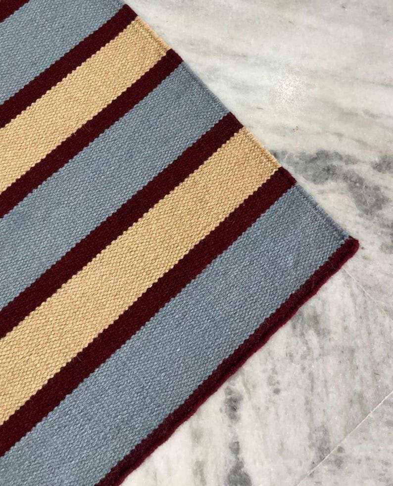 May include: A close-up of a striped rug with alternating bands of light blue, beige, and burgundy. The rug's texture is visible, and the color scheme creates a visually appealing pattern. The rug is likely made of woven material.