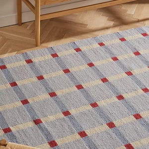 Pode incluir: Um tapete tecido azul claro e creme com um padrão xadrez de quadrados vermelhos. O tapete é retangular e parece ser feito de fibras naturais. O tapete está em um piso de madeira.