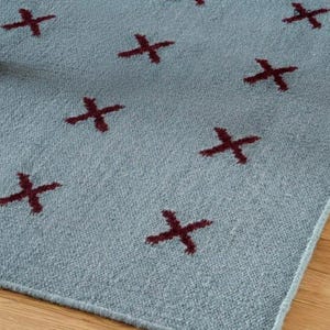 Könnte beinhalten: Ein hellblauer Teppich mit einem sich wiederholenden Muster aus dunkelroten Kreuzsymbolen. Der Teppich hat eine texturierte, gewebte Optik mit einer gleichmäßigen, gitterartigen Anordnung der kleinen, dunkelroten X-förmigen Motive.