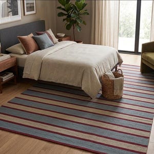 May include: A bedroom scene featuring a bed with a gray headboard, pillows, and a beige comforter. A striped rug in shades of blue, beige, and burgundy covers the floor. A woven basket with a blanket sits near the bed, and a green armchair is in the background.