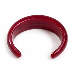 Peut inclure: Un bracelet jonc rigide en forme de C, d'une couleur rouge profond avec une finition lisse, polie et brillante. Le bracelet présente un profil épais et arrondi ainsi qu'une large ouverture, affichant une teinte cramoisie uniforme.