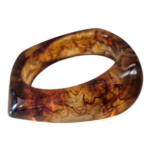 Puede incluir: Un anillo translúcido de color ámbar con una forma cuadrada redondeada única. El interior del anillo presenta patrones arremolinados de marrón oscuro y ámbar, creando un efecto veteado. El anillo es un accesorio de moda.