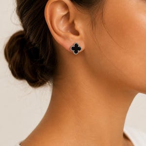 May include: A stud earring featuring a black four-leaf clover shape with a metallic beaded border. The clover design is centered on the earlobe, showcasing a smooth, dark, opaque inlay surrounded by a textured, reflective silver-toned metal edge.