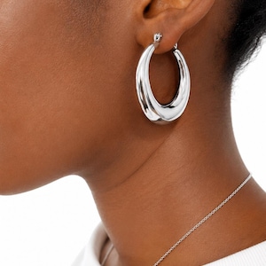May include: A polished silver-tone hoop earring with a thick, rounded, and tapered design. The earring features a smooth, reflective metallic surface and a secure hinged closure mechanism that connects at the top of the circular hoop structure.